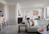 A modern living room with a sofa, coffee table, and chairs. There is a dining area with a table and chairs. A chandelier hangs from the ceiling, and a painting is on the wall.