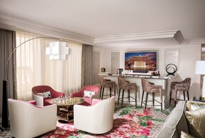 A hotel room with a bar area, two pink armchairs, a round table, and a large floral carpet.