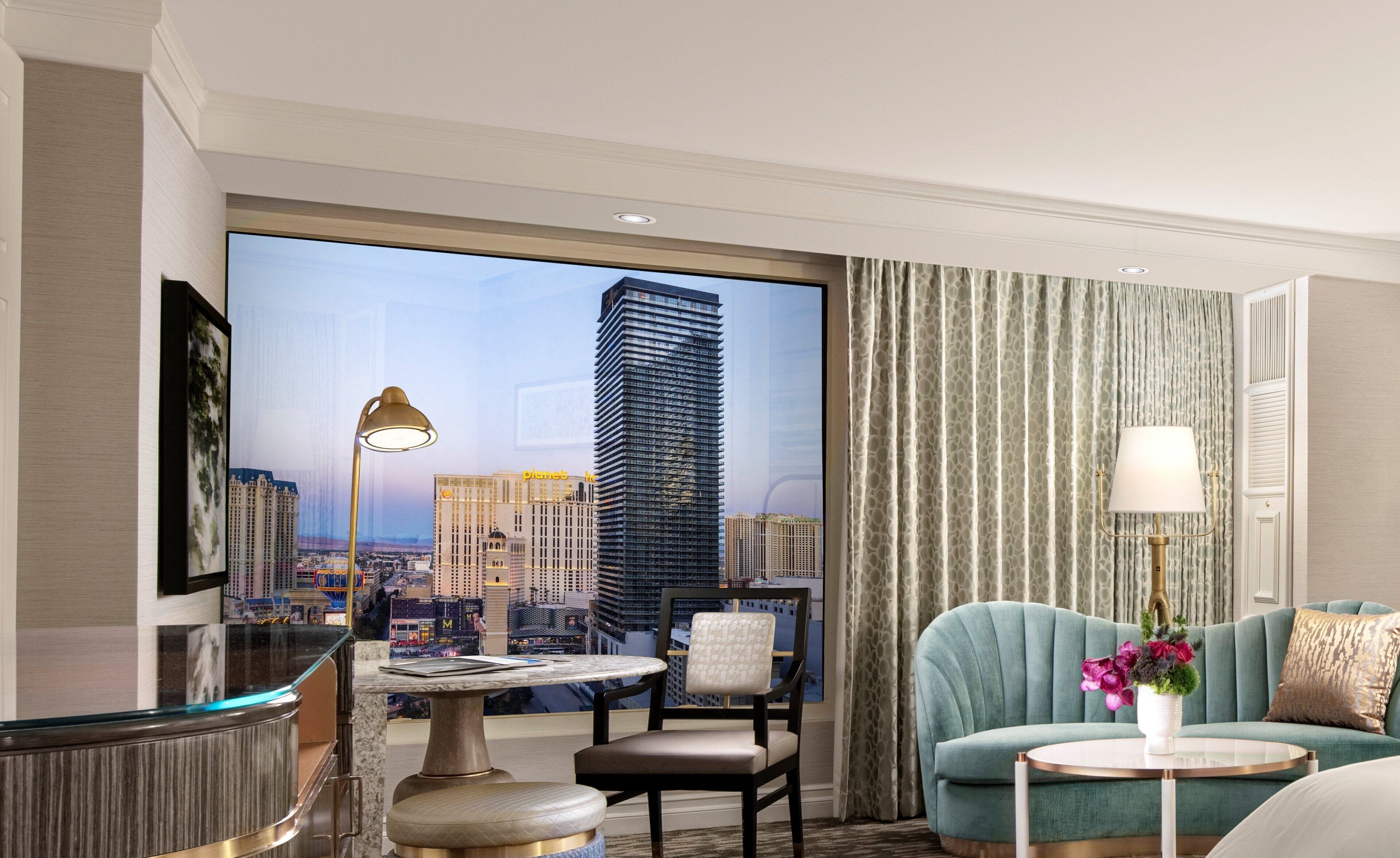A hotel room with a large window offering a city view, a round table, a sofa, and a lamp.
