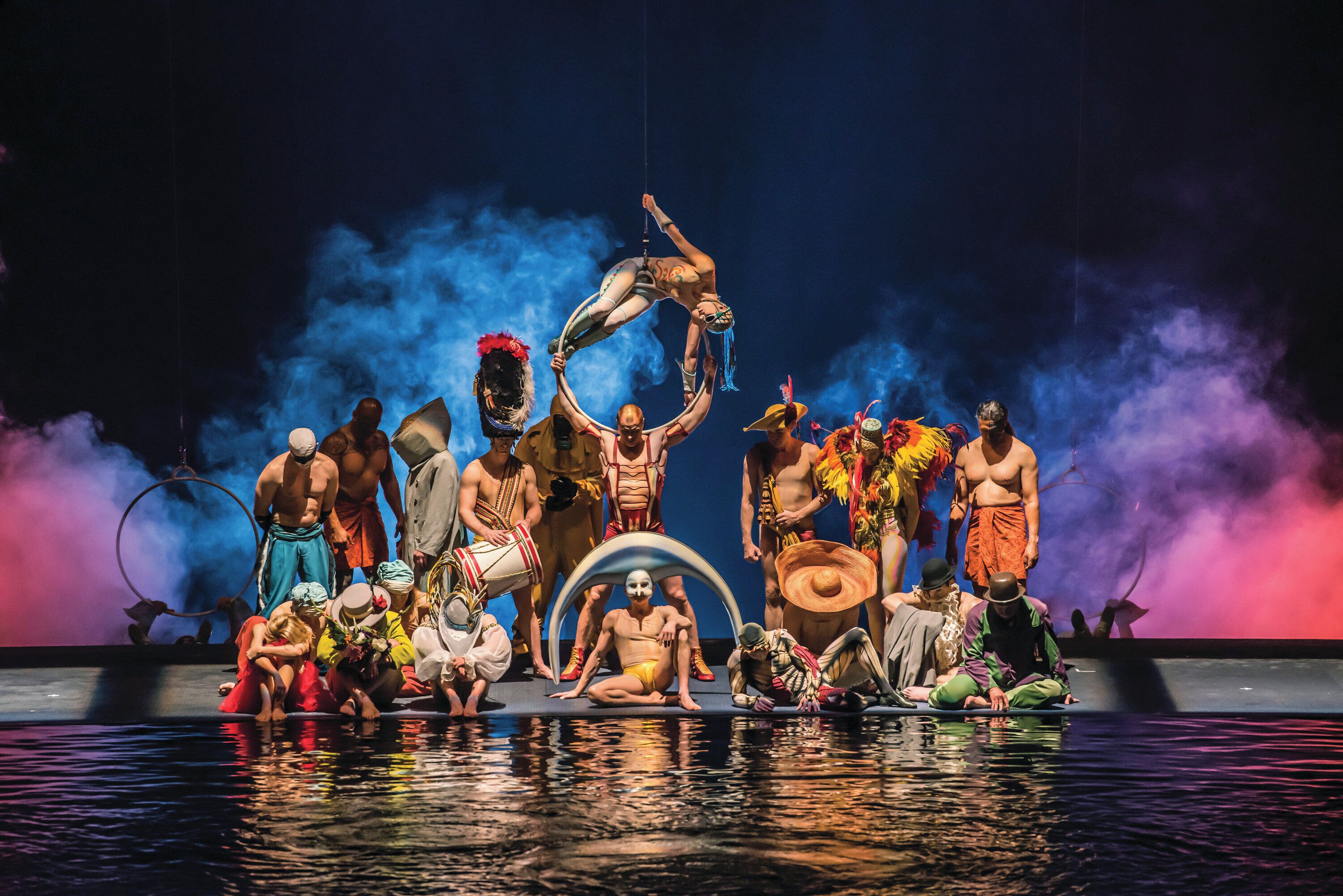 A group of people in various costumes on a stage with a reflective water surface.