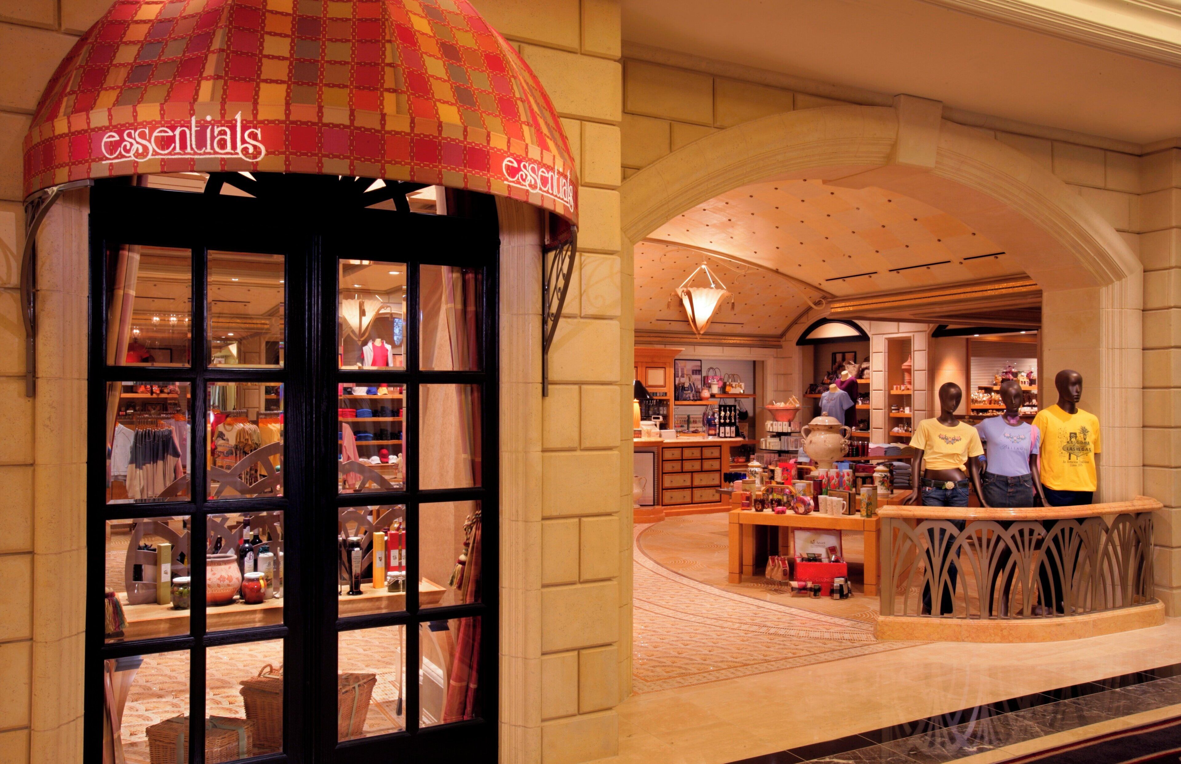 A store named 'essentials' with a checkered awning and a display window. Inside, there are shelves with various items and a counter with a person standing behind it.