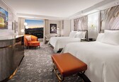 A hotel room with two beds, a desk, a chair, and a view of the cityscape.