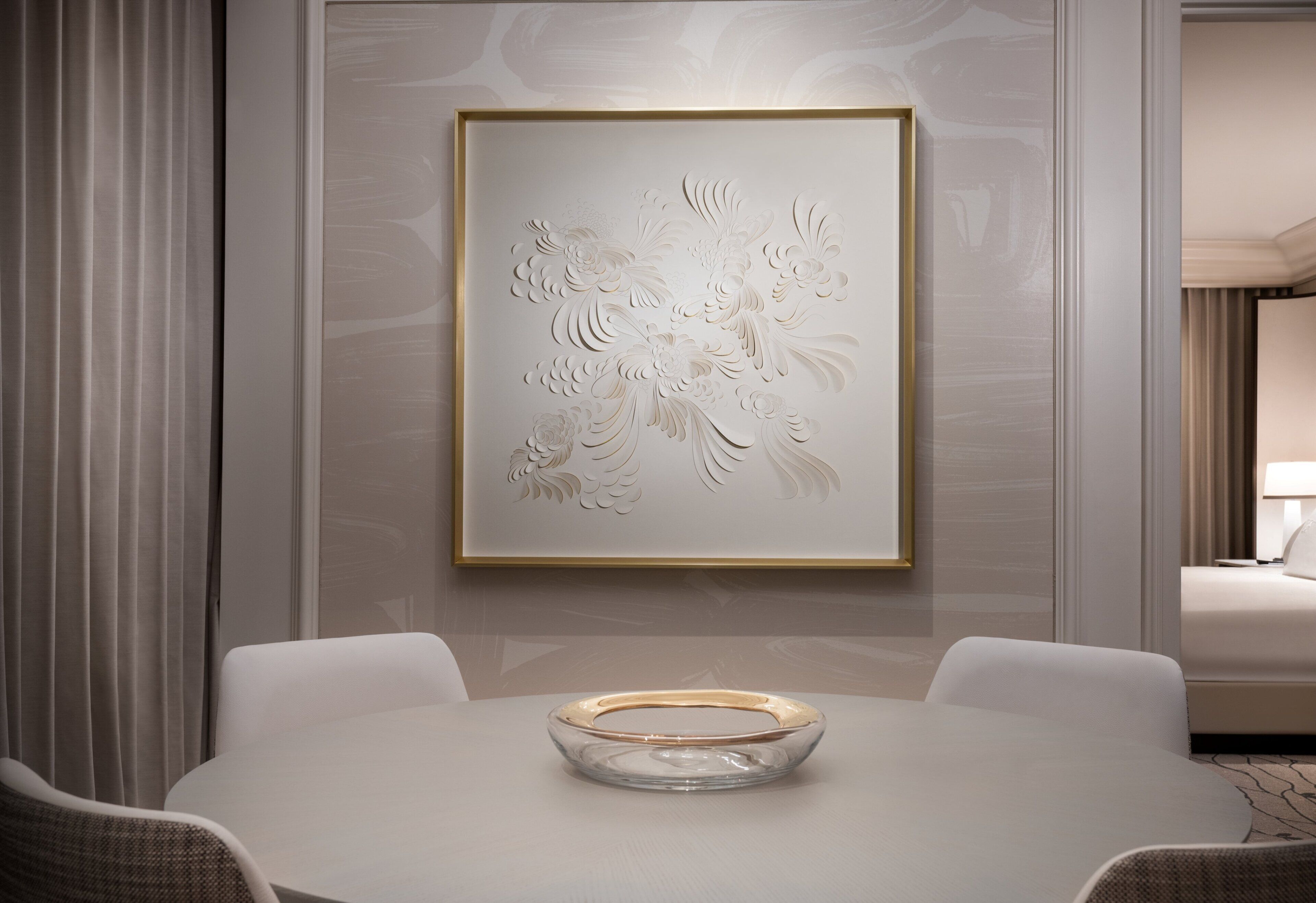 A modern hotel room with a dining area featuring a white table, a clear glass bowl centerpiece, and a framed floral artwork on the wall.