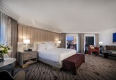 A hotel room with a large bed, a desk with a lamp, a seating area with a sofa and armchair, and a view of the city through the window.
