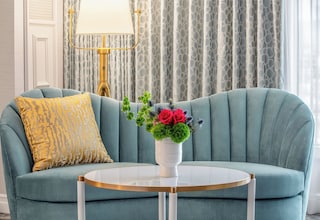 A teal sofa with a patterned yellow pillow, a white vase with flowers, and a gold lamp.