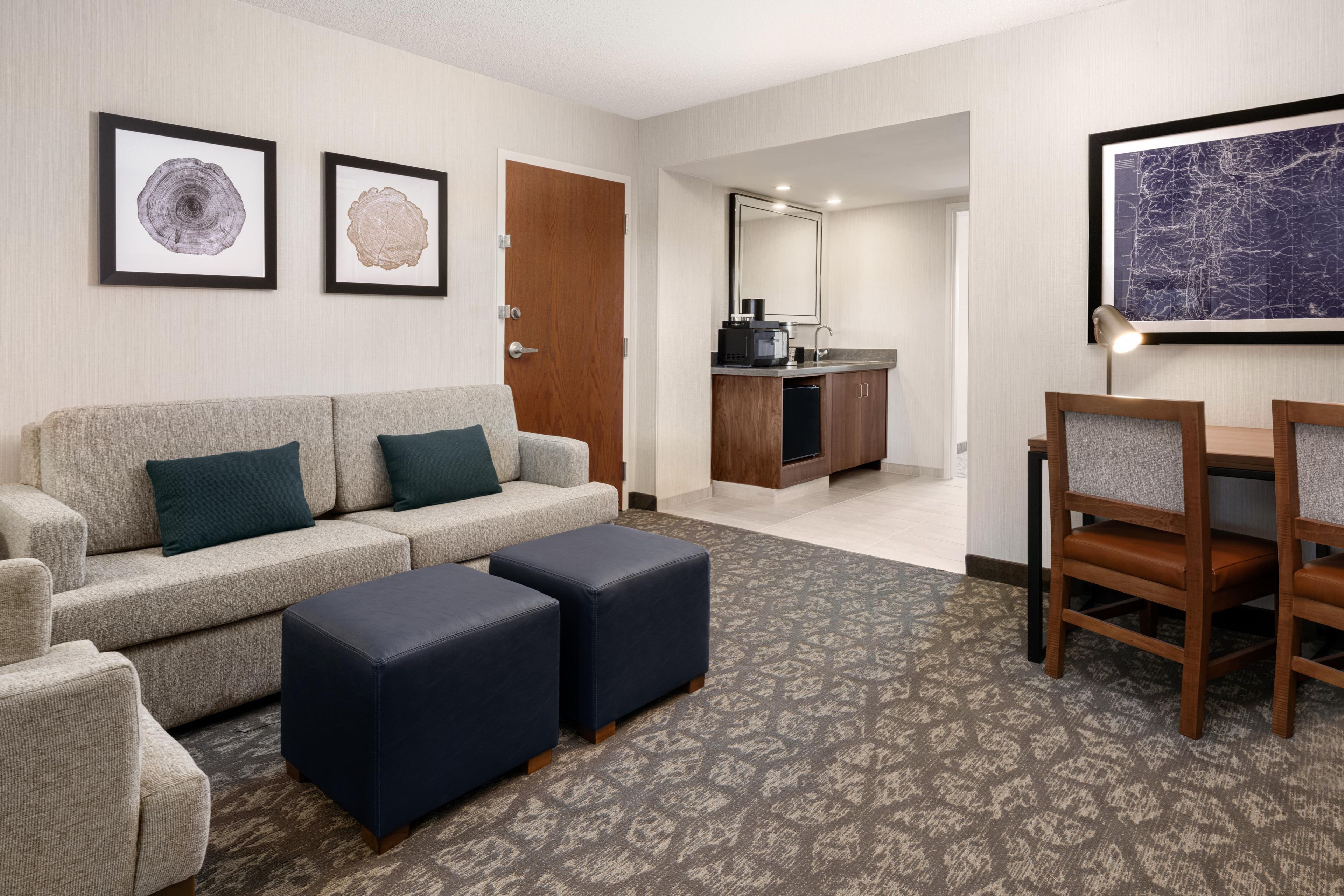 A hotel room with a sofa, ottomans, a coffee machine, a dining table with chairs, and artwork on the walls.
