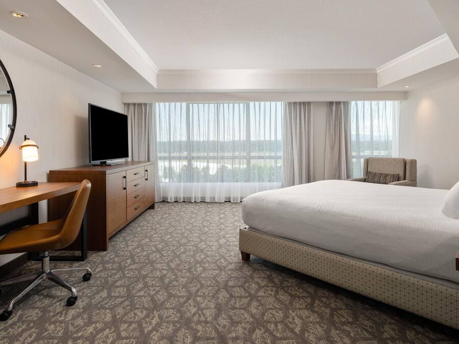 Una camera d'albergo con un letto, una scrivania, una sedia, una televisione e un'ampia finestra con tende.