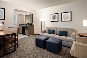 A hotel room with a dining table, a sofa, two ottomans, a lamp, and a kitchenette.