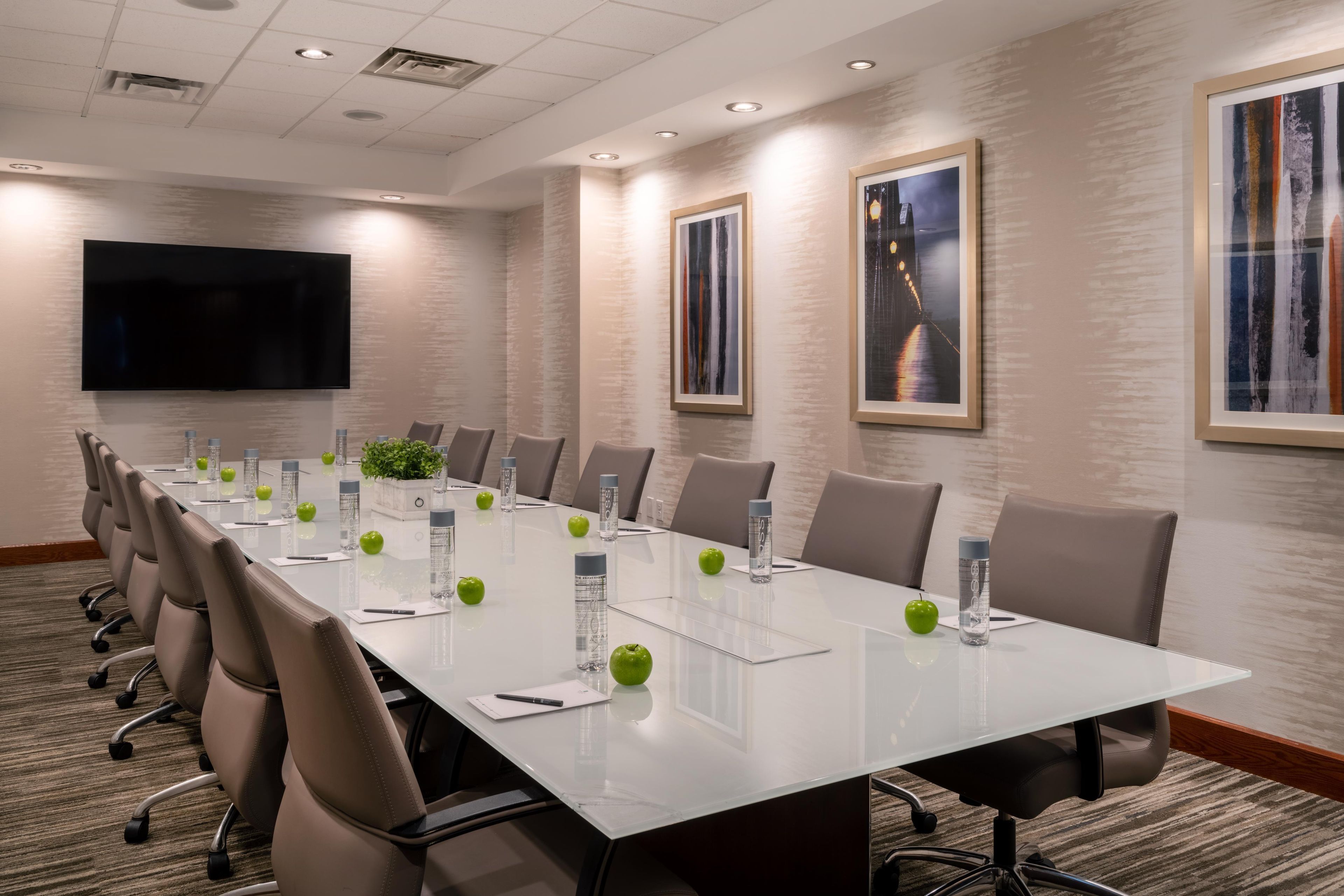 A conference room with a long table, chairs, a flat-screen TV, framed artwork, and a plant.