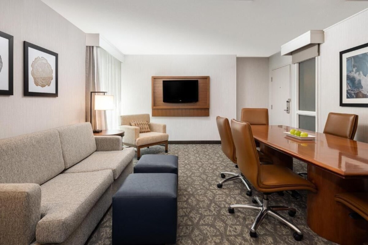 A hotel room with a sofa, armchairs, a desk with chairs, a flat-screen TV, and framed artwork on the walls.