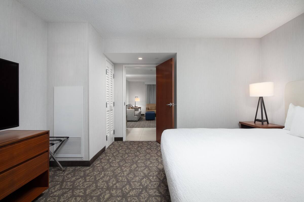 Habitación de hotel con cama, televisor, cómoda, lámpara y vista hacia la habitación contigua.