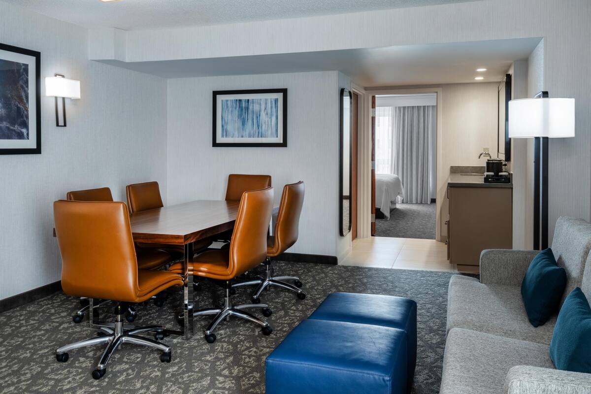 Habitación de hotel con una mesa de conferencias, sillas de cuero, un sofá, un reposapiés azul, una lámpara y un cuadro enmarcado en la pared.