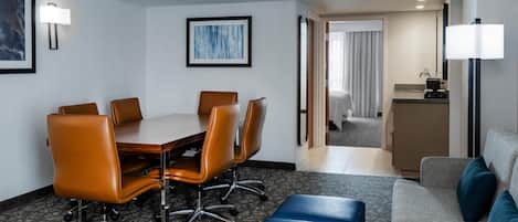 Habitación de hotel con una mesa de conferencias, sillas de cuero, un sofá, un reposapiés azul, una lámpara y un cuadro enmarcado en la pared.