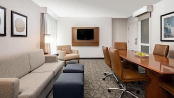 A hotel room with a sofa, two armchairs, a coffee table, a TV mounted on the wall, and a conference table with chairs.