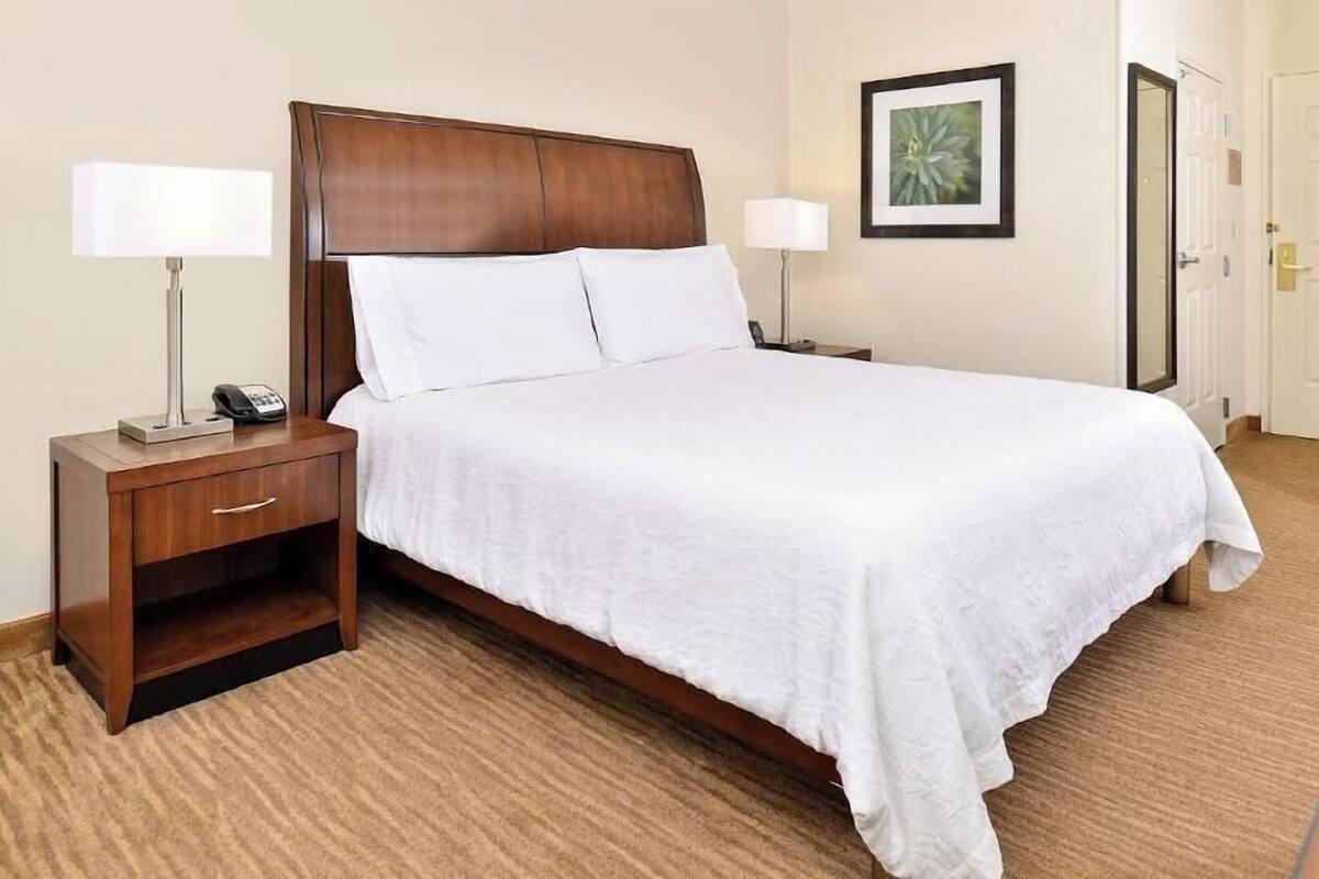 Una habitación de hotel con una cama grande, dos mesitas de noche con lámparas, una mesilla auxiliar y un cuadro en la pared, además de una puerta.