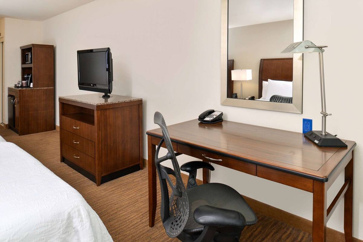 Habitación de hotel con cama, escritorio con monitor de computadora, teléfono, silla, lámpara de escritorio y espejo.