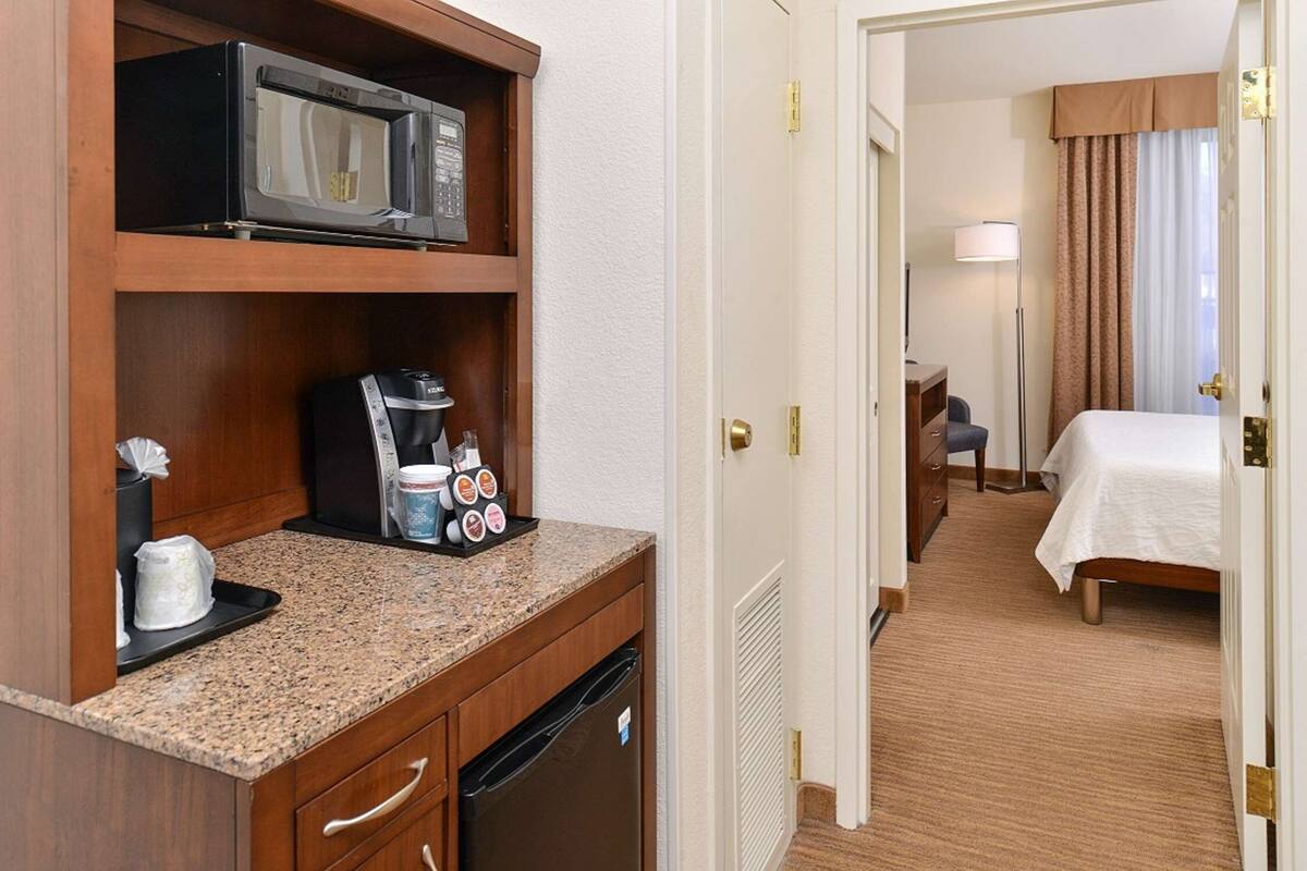 Habitación de hotel con kitchenette, que incluye microondas, cafetera y refrigerador. Hay una cama con cubrecama blanco y una lámpara.