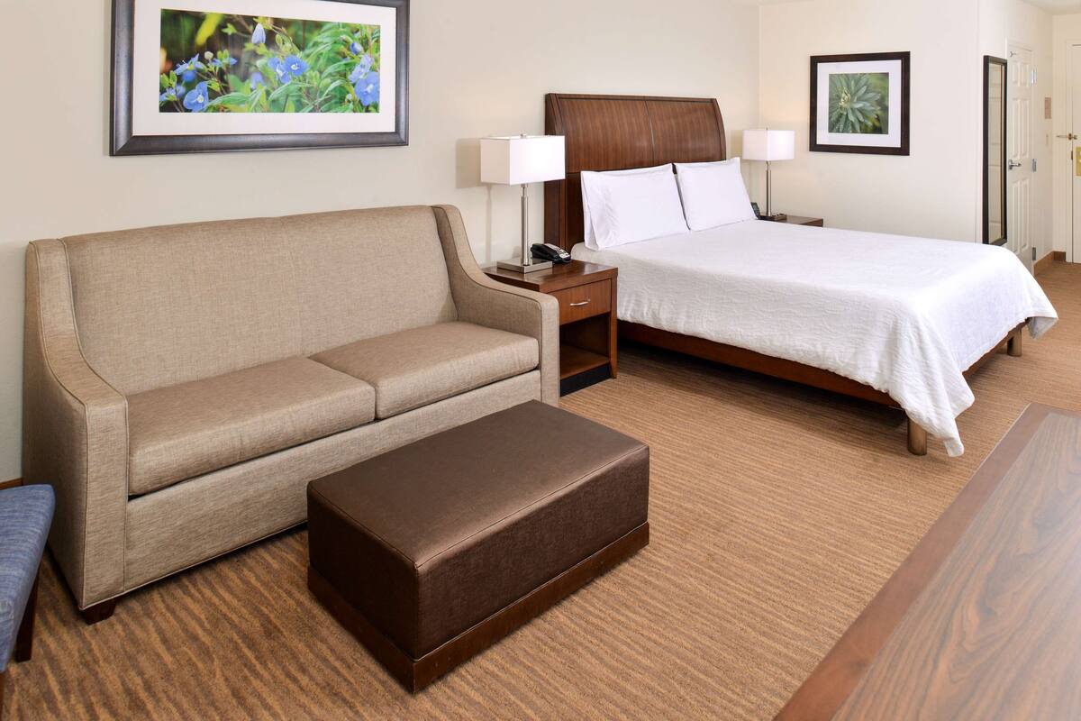 Habitación de hotel con sofá color crema, cama de madera con ropa de cama blanca, mesita de noche con lámpara y un cuadro en la pared.