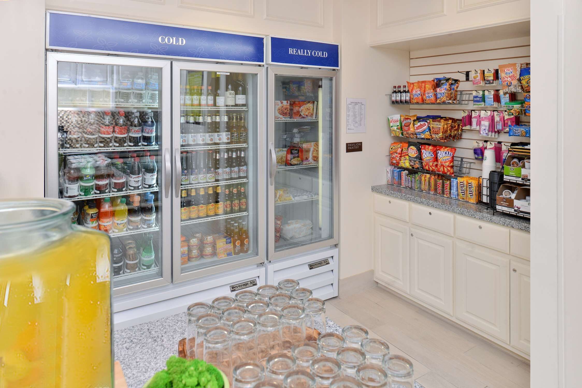 A kitchen area with two beverage refrigerators labeled "COLD" and "REALLY COLD," stocked with various drinks. There are also shelves with snacks and condiments.