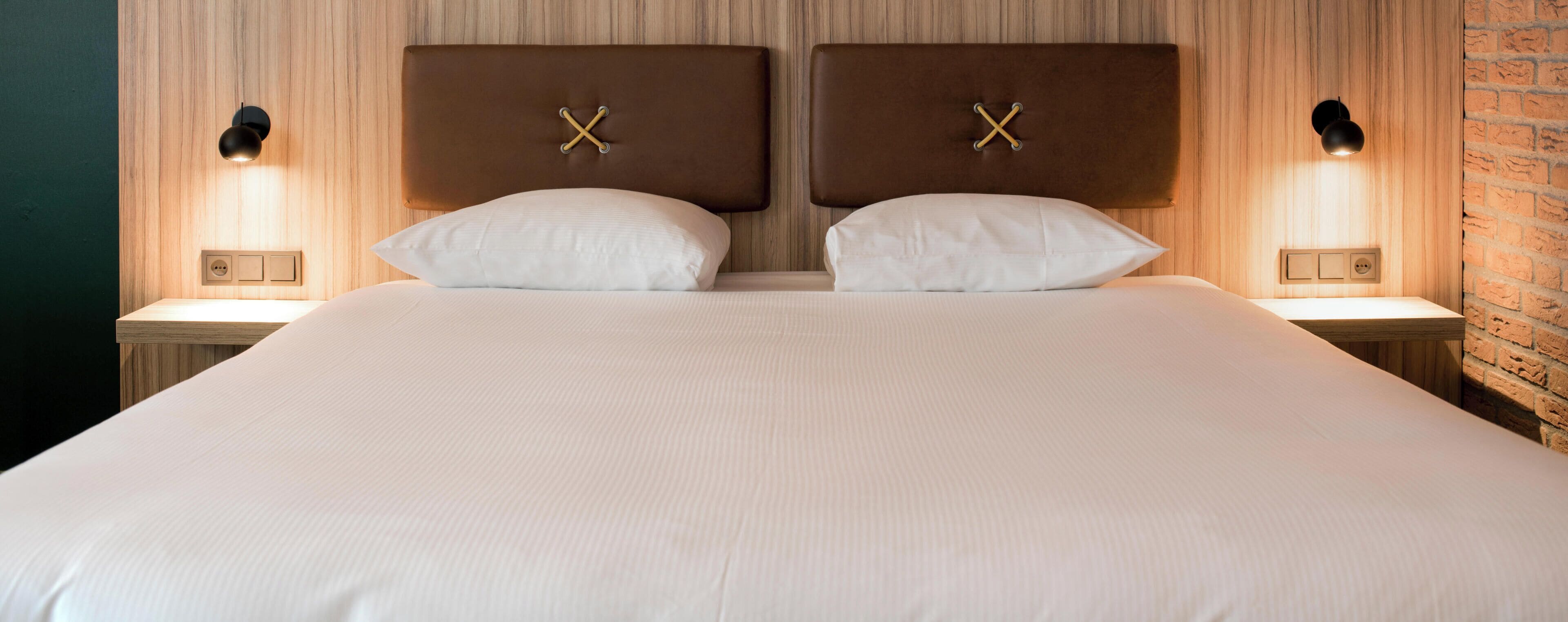 Een tweepersoonsbed met hoofdbord met een kruisdecoratie, met aan weerszijden wandlampen, in een kamer met houten lambrisering en een accentmuur van baksteen.