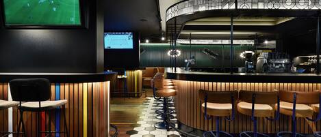 A bar area with a curved counter, multiple stools, and a large screen displaying a sports game.