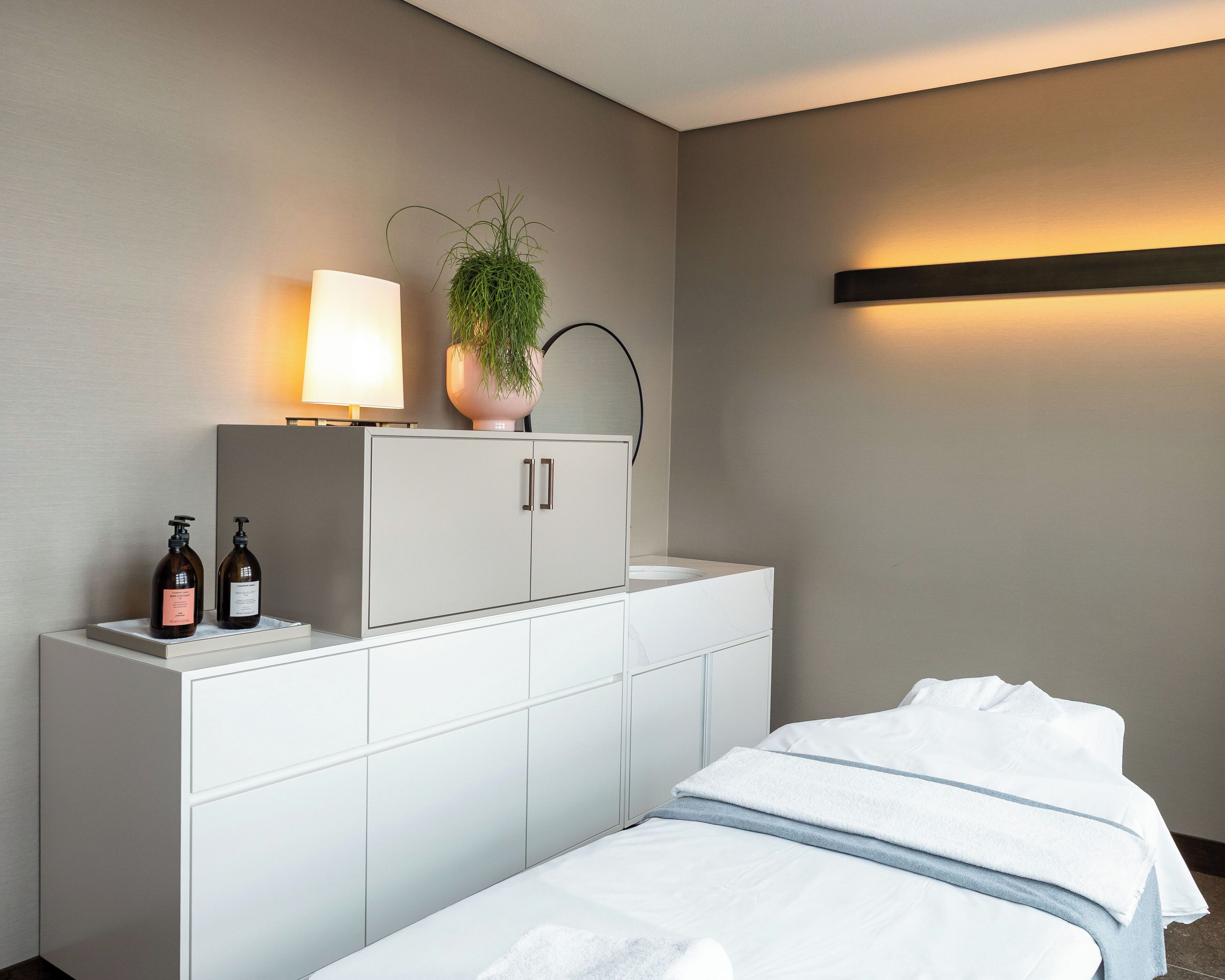 Una habitación de hotel con una cama, un armario blanco con dos botellas, una planta en maceta y una lámpara fijada a la pared.