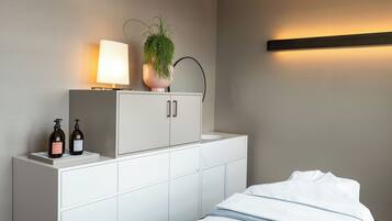 Una habitación de hotel con una cama, un armario blanco con dos botellas, una planta en maceta y una lámpara fijada a la pared.