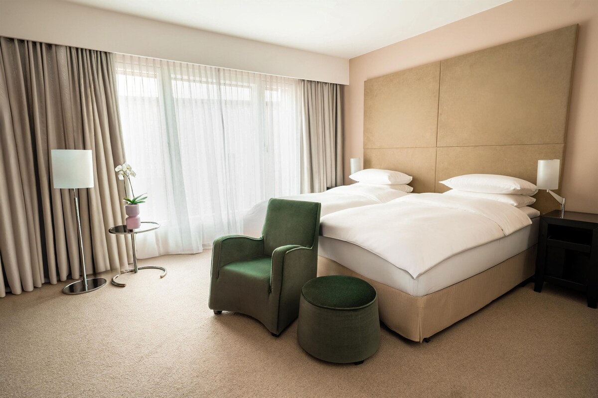 A hotel room with a large bed, a green armchair, a small table with a lamp, and a potted plant.