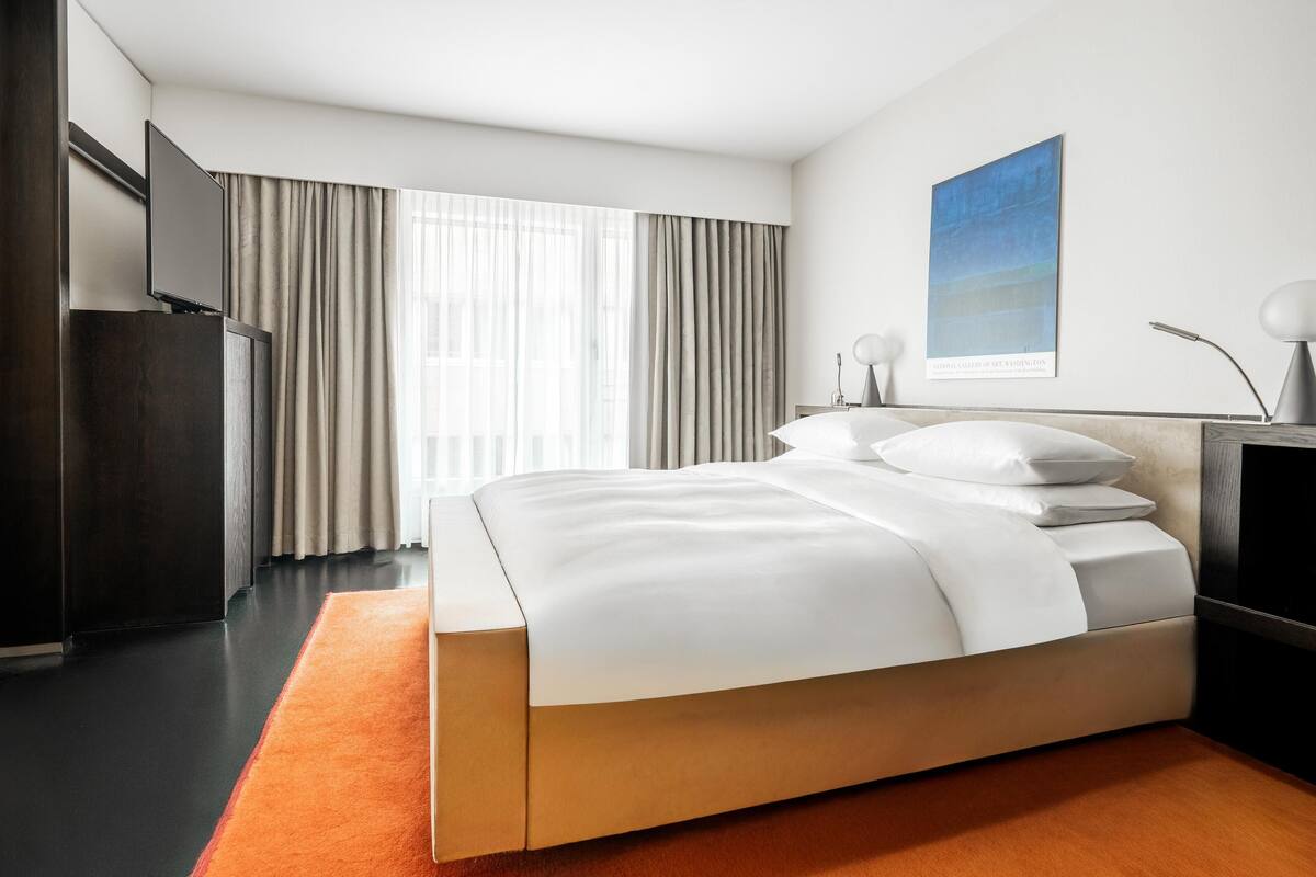 A modern hotel room with a large bed, a desk, a television, and a window with curtains.