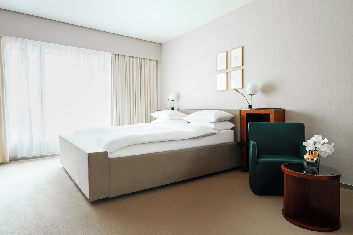 A modern hotel room with a large bed, a green armchair, a small wooden side table, and a large window with sheer curtains.