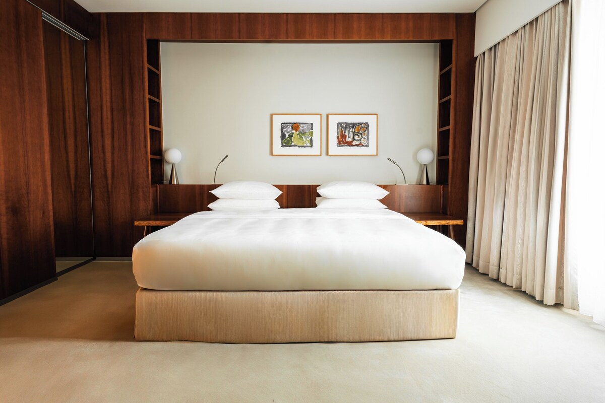 A modern bedroom with a large bed, wooden paneling, a bedside table, two framed artworks, and a large window with curtains.