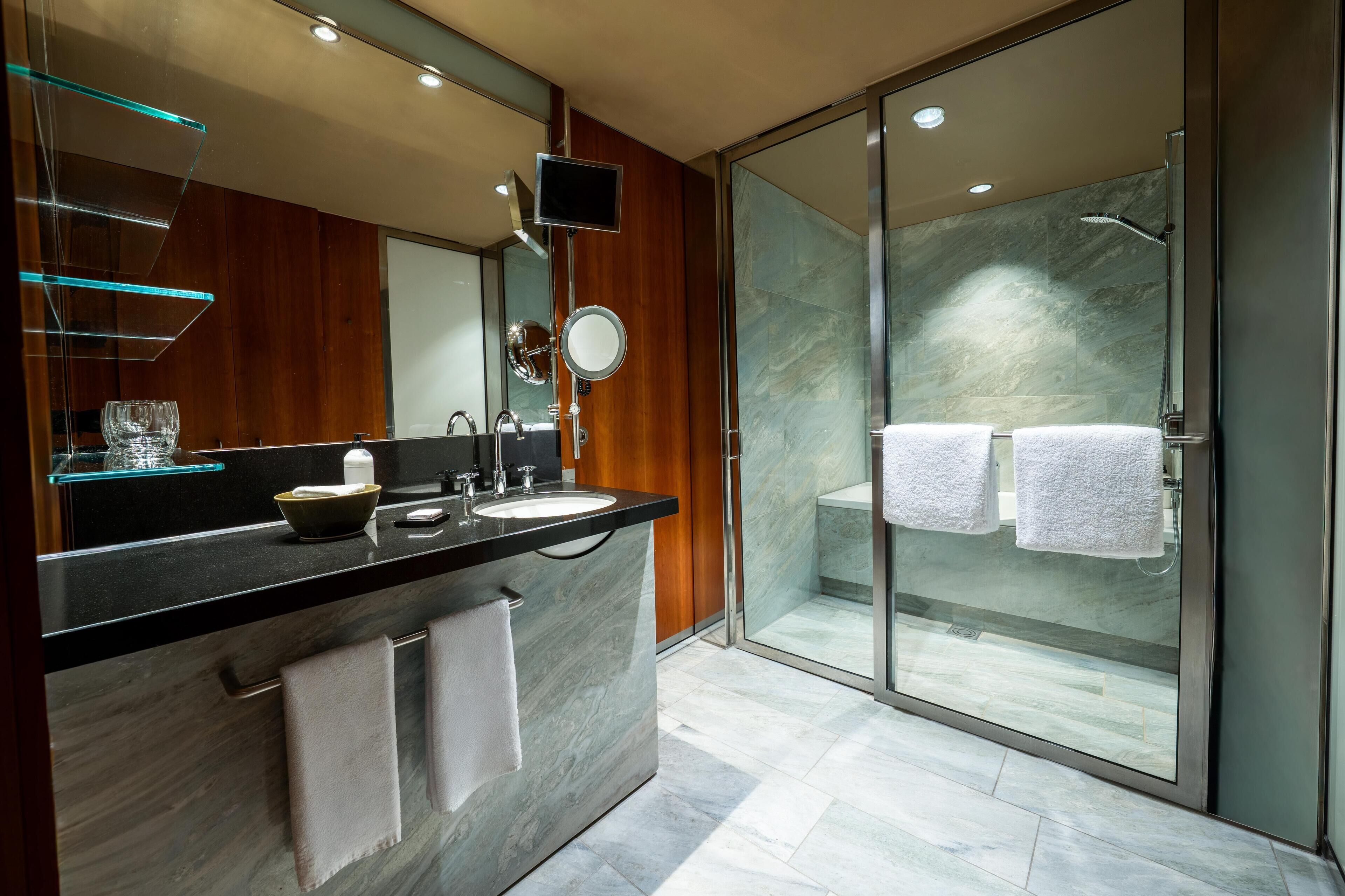 A modern bathroom with a glass-enclosed shower, two sinks, a large mirror, and a glass shelf with a bowl and glass.