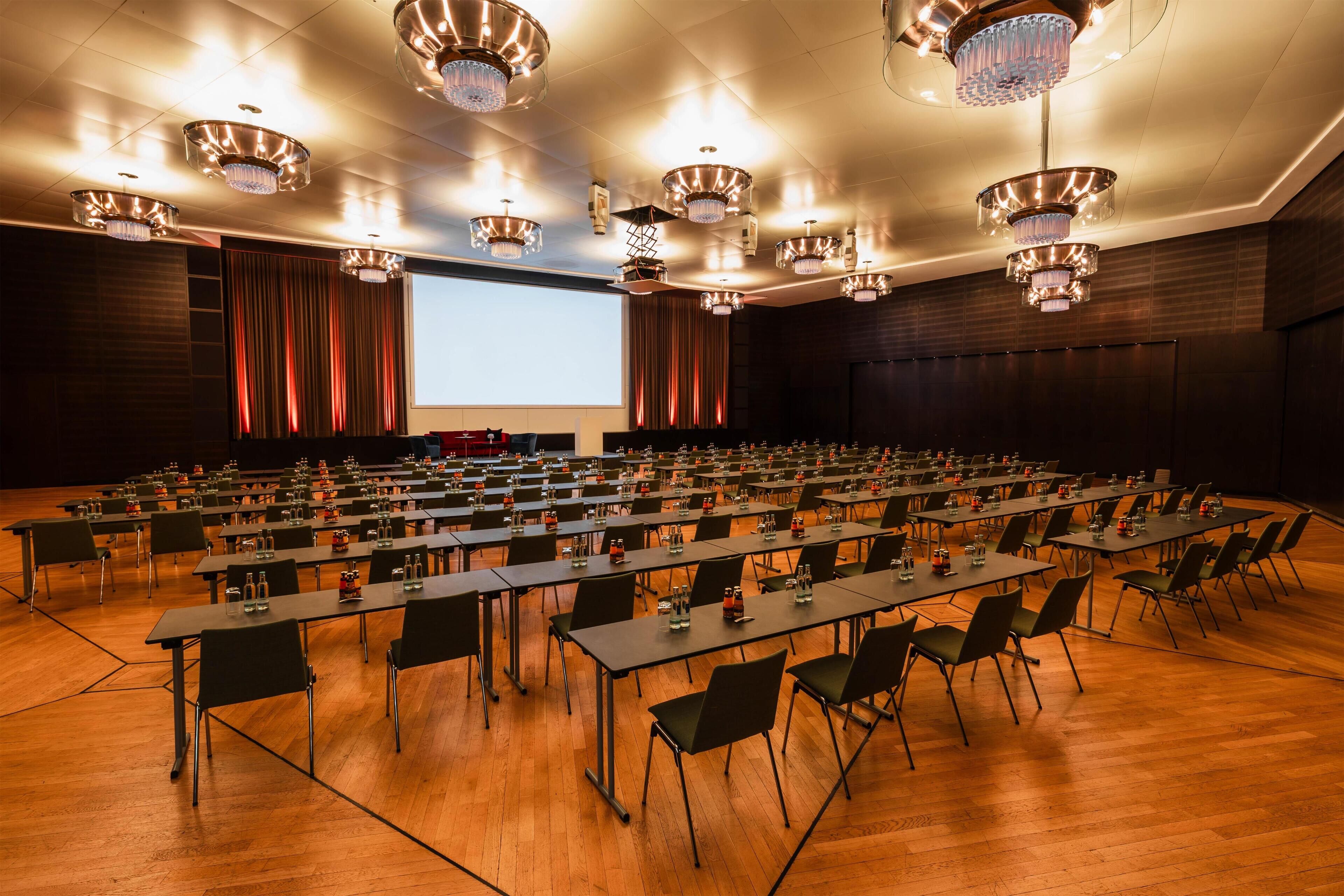 A conference room with rows of tables and chairs, large chandeliers, and a screen at the front.