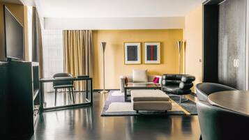 A modern living room with a sofa, ottoman, and a TV. There are two framed pictures on the wall, a floor lamp, and a large window with curtains.