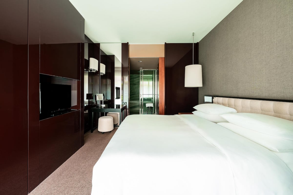 A modern hotel room with a large bed, a flat-screen TV, a desk with a chair, and a view of the hallway.