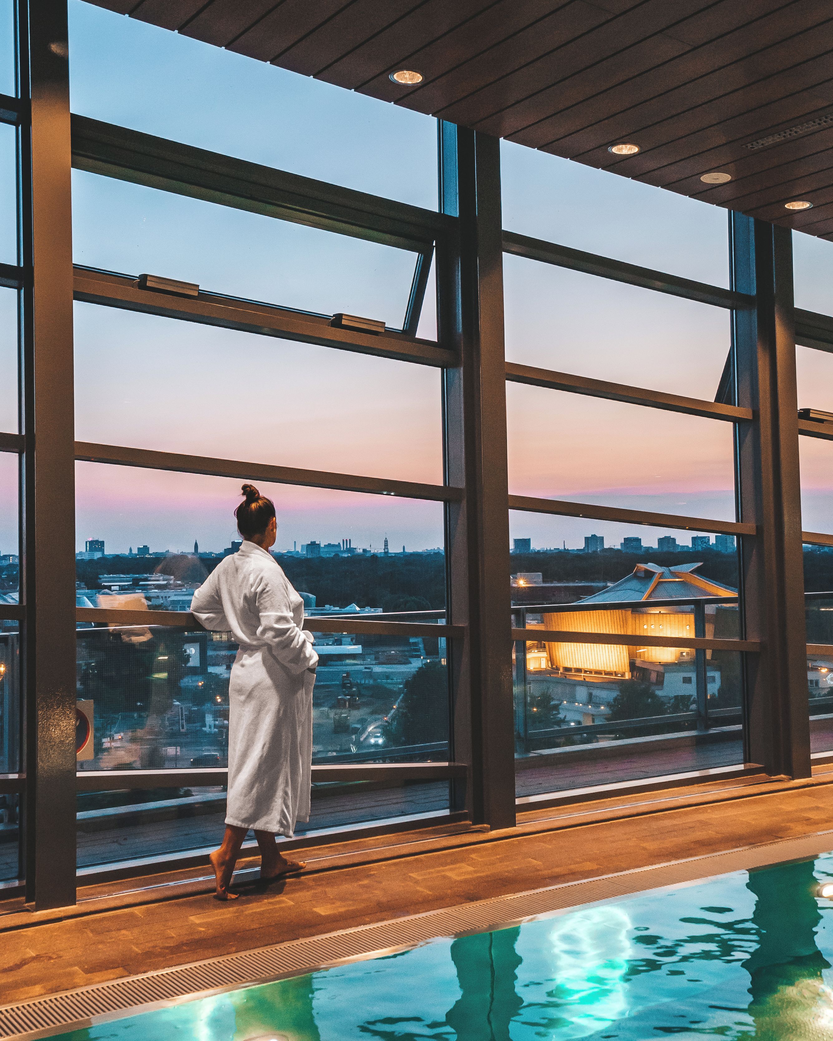 Una persona en bata de baño de pie junto a una amplia piscina interior con vistas a la ciudad a través de grandes ventanales.