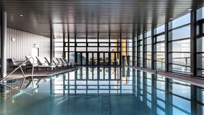 An indoor swimming pool with clear glass walls, lounge chairs, and a view of the cityscape. - Grand Hyatt Berlin (Berlin)