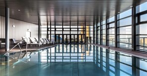 Una piscina interior con paredes de vidrio transparente, sillones de descanso y vistas a la ciudad.