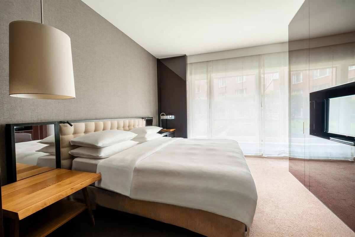 A modern hotel room with a large bed, a wooden nightstand, a large window with sheer curtains, and a flat-screen TV.