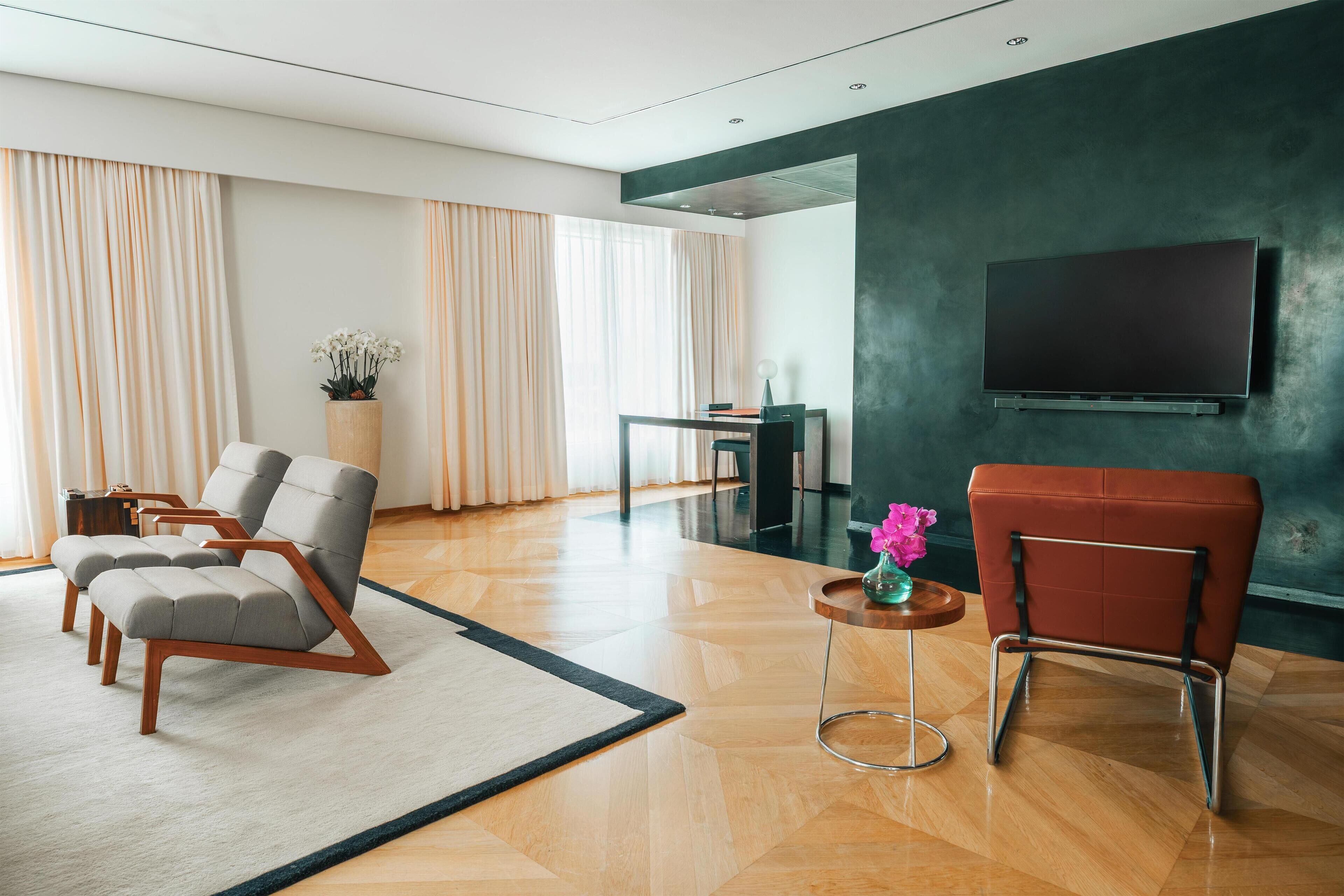 Una sala de estar moderna con un televisor de pantalla plana, dos sillones grises, un sillón rojo, una mesa de centro de madera y una planta en maceta.