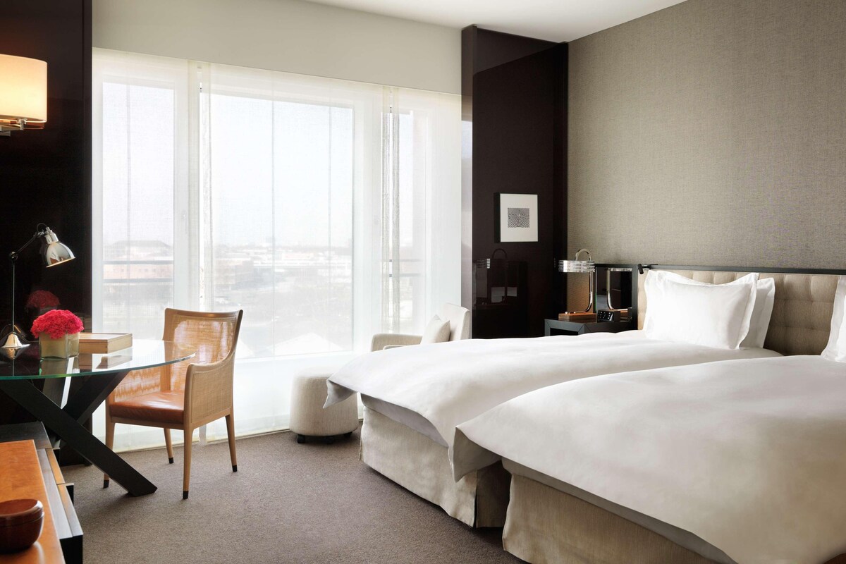 A hotel room with two beds, a desk, a chair, and a large window with a city view.
