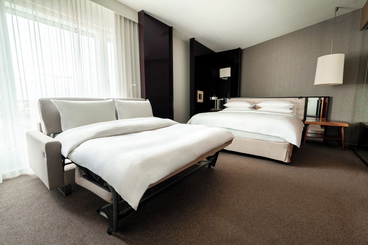 A modern hotel room with two beds, a sofa, a desk, and a large window with sheer curtains.