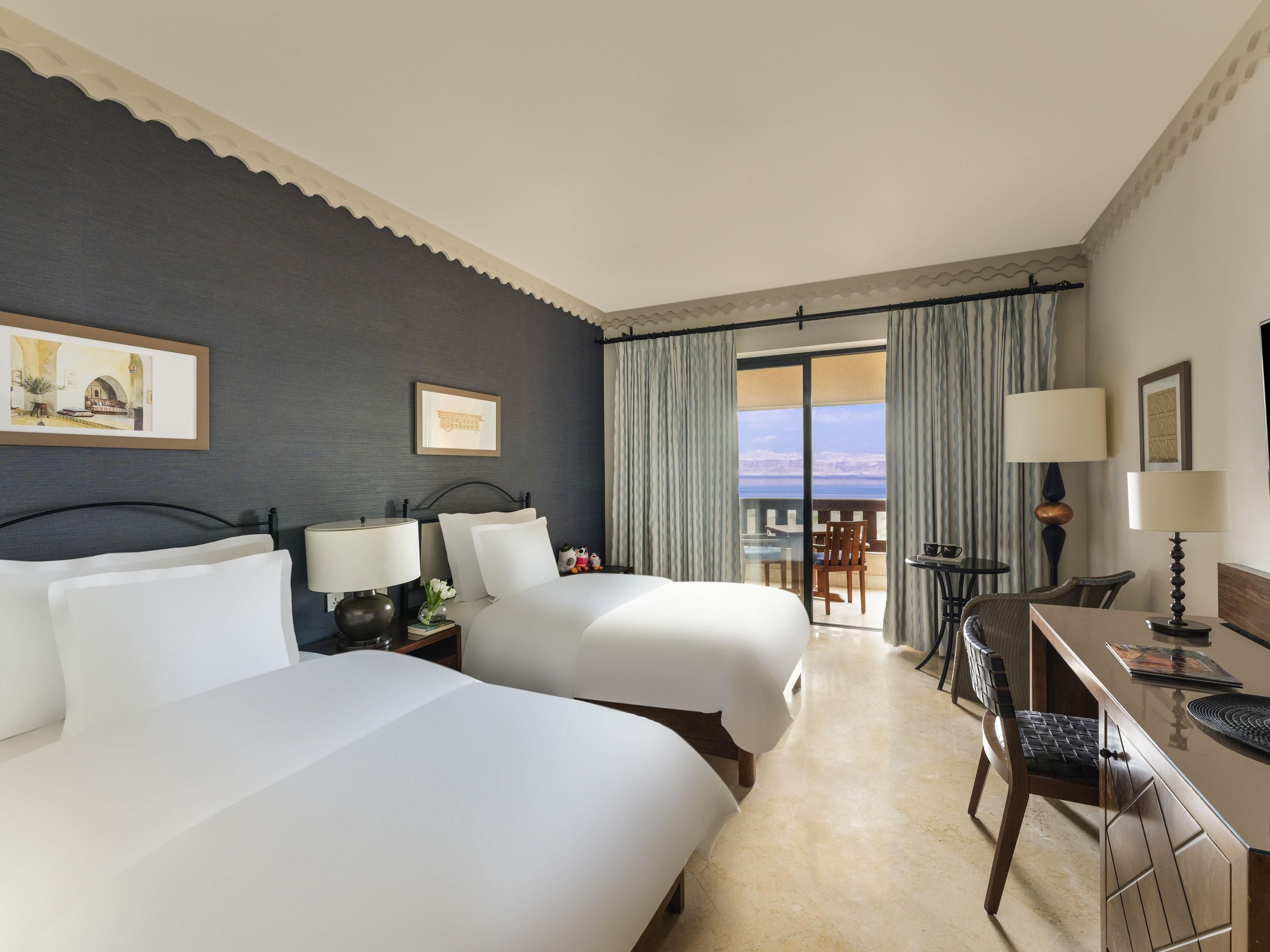 A hotel room with two beds, a desk, a chair, and a balcony with a view.