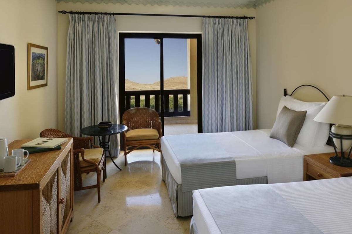 A hotel room with two beds, a desk with a chair, a small table, and a balcony with a view.