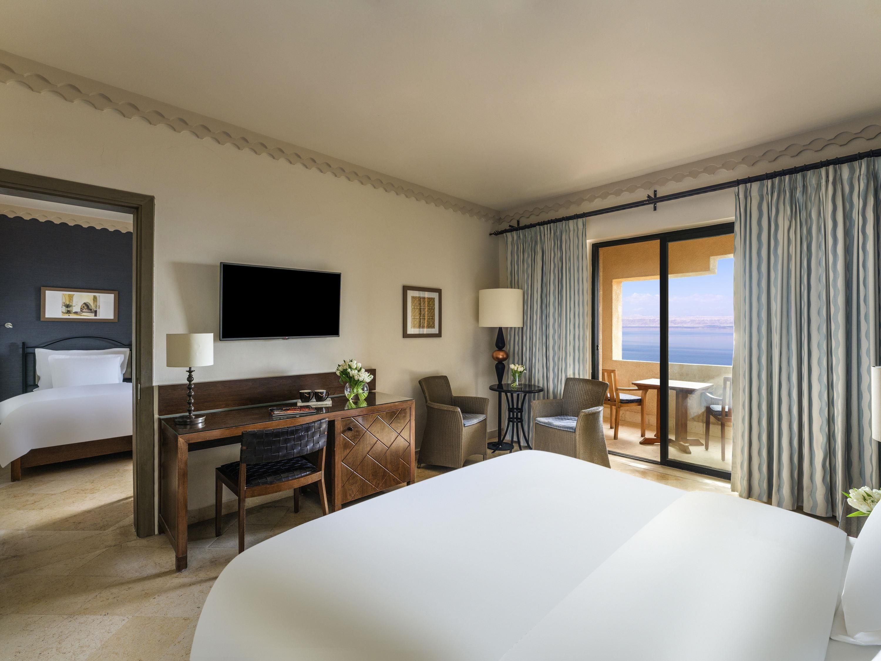 A hotel room with a large bed, a desk with a chair, a television, and a view of the sea through the window.