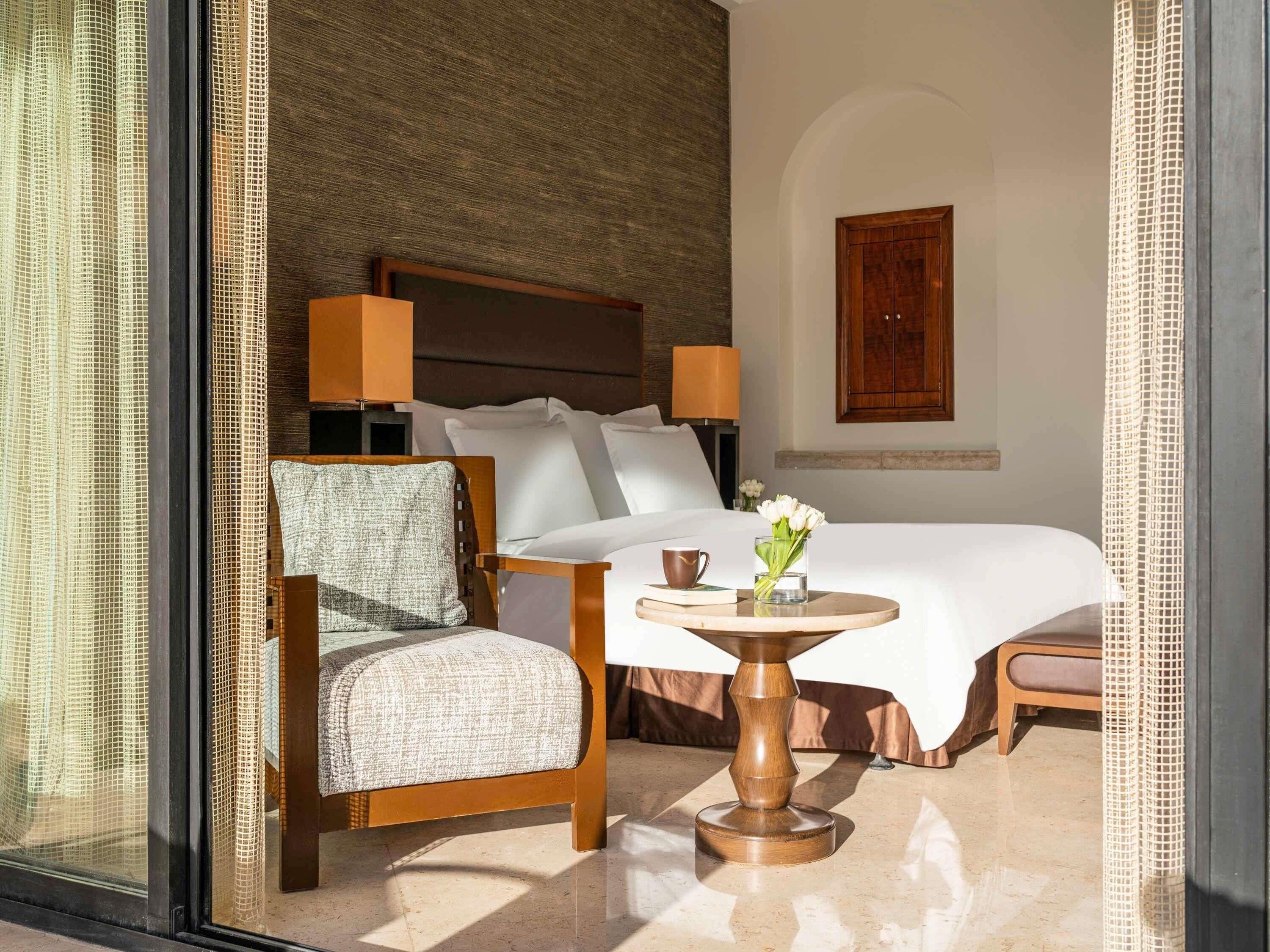 A hotel room with a bed, a chair, a table with a vase of flowers, and a lamp.