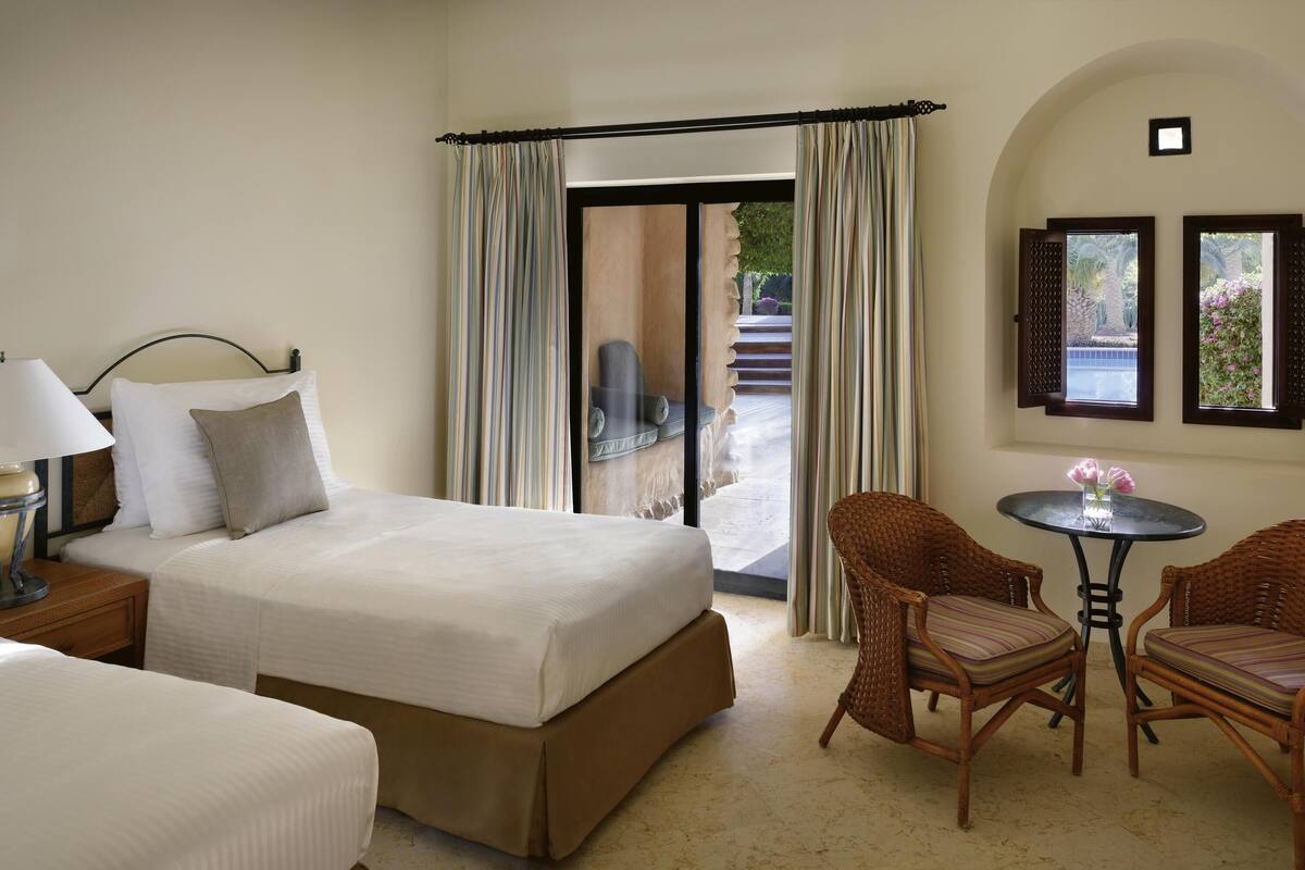 A hotel room with a bed, a nightstand, a lamp, a chair, a table with flowers, and a view of the outdoors.