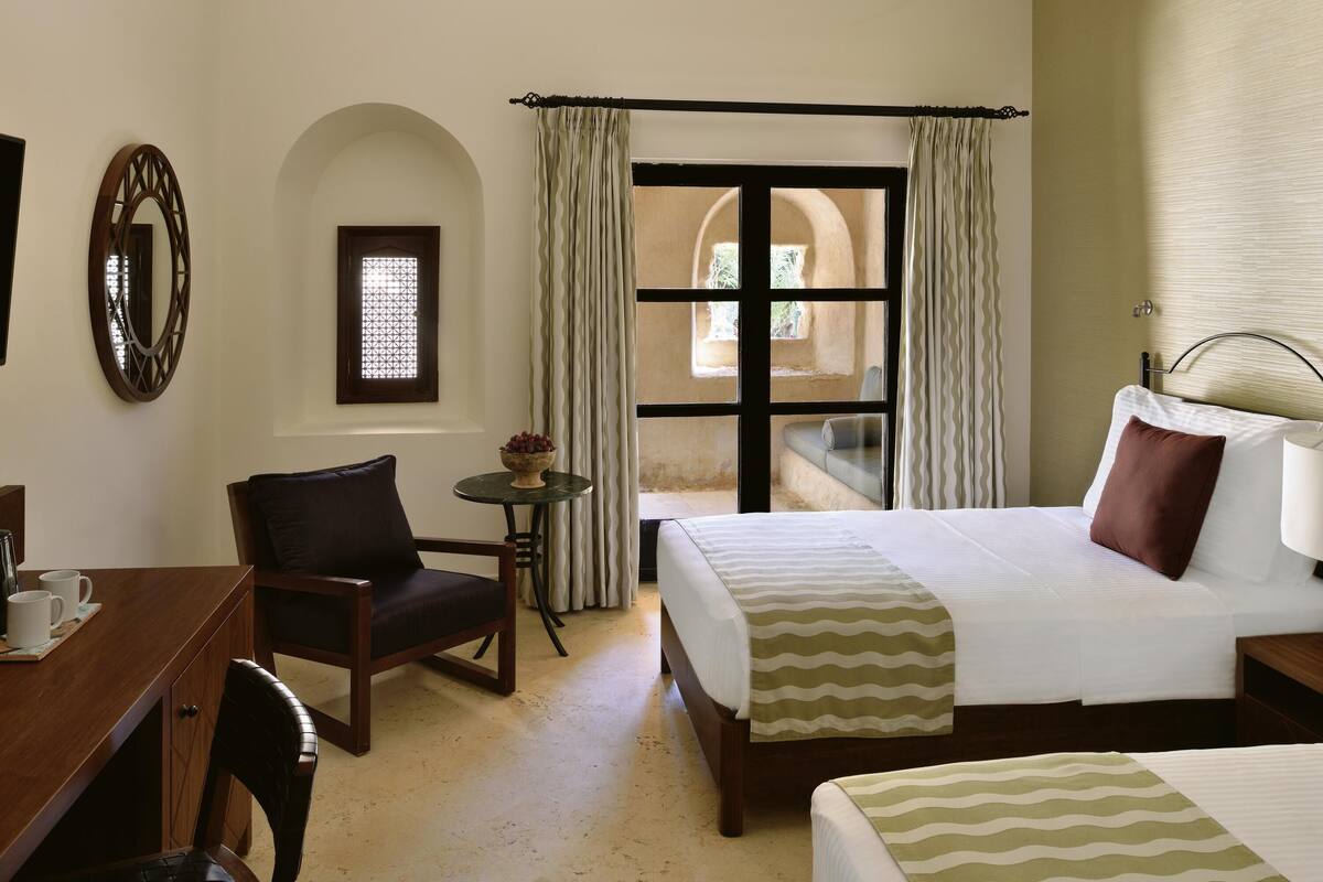 A hotel room with a bed, a chair, a small table with a bowl of fruit, a window with curtains, and a view of the outside.