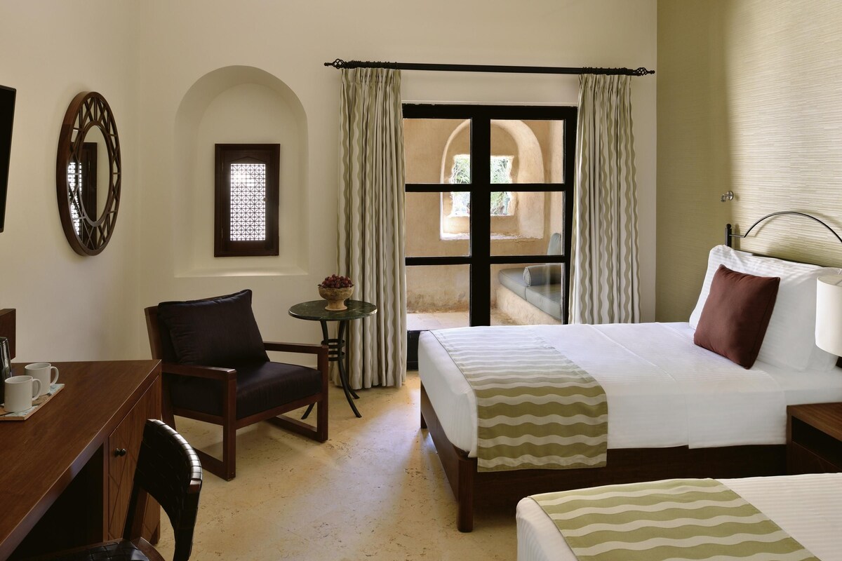 A hotel room with a bed, a chair, a small table with a bowl of fruit, and a window with curtains.