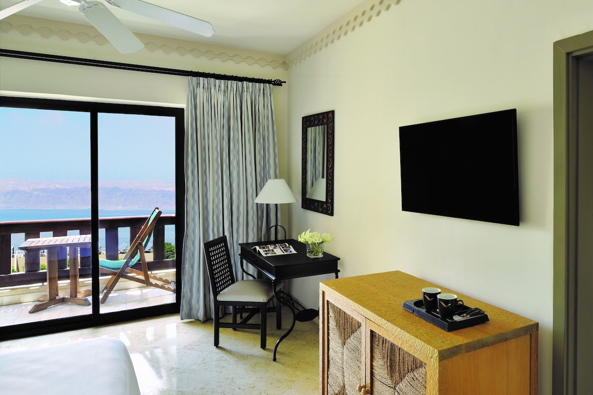 A room with a balcony, a desk with a lamp, a chair, a television, and a small table with cups.
